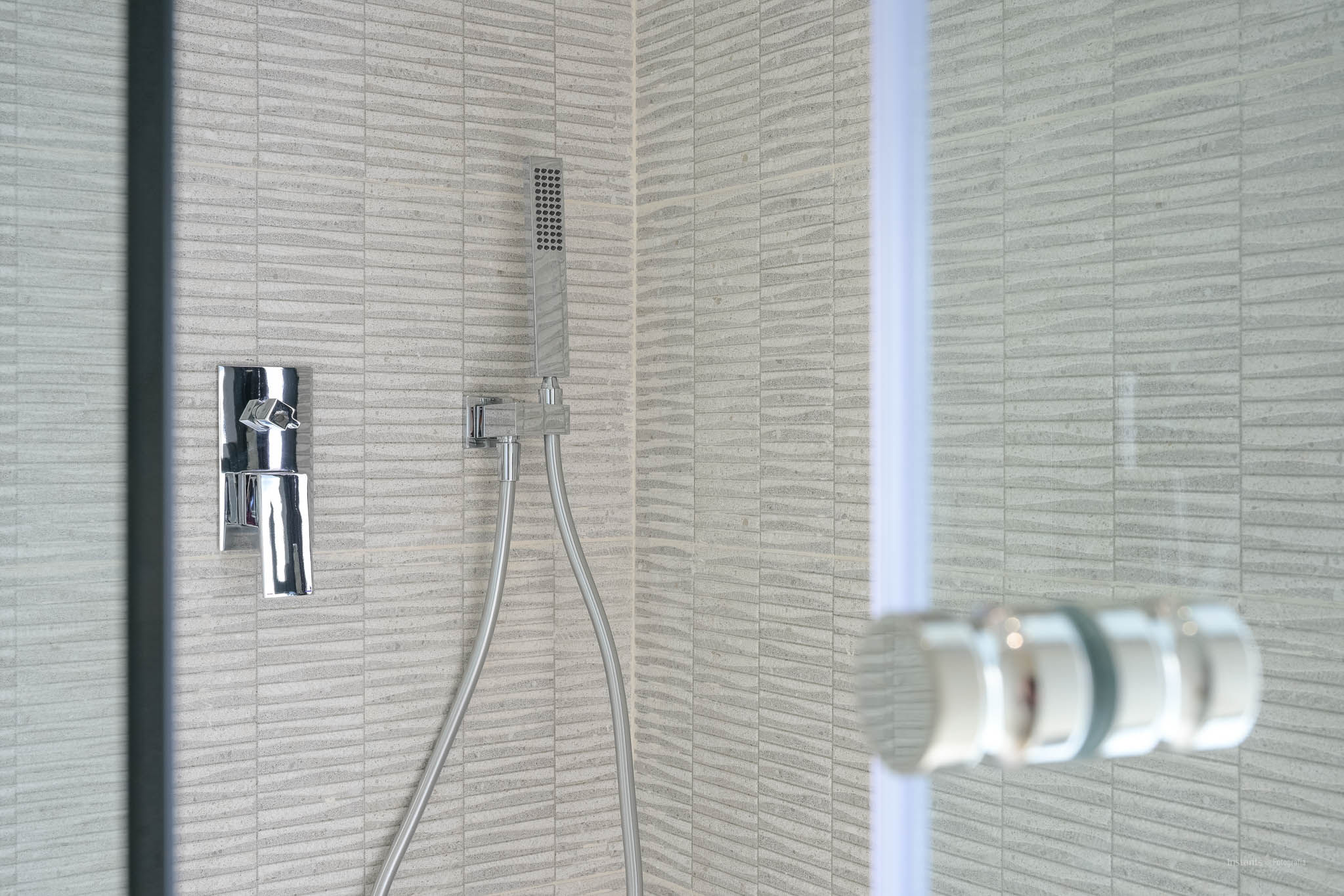 Detail van een moderne douche met grijze, gestructureerde rechthoekige tegels, een slanke chromen mengkraan en handdouche. Een glazen scherm is zichtbaar.