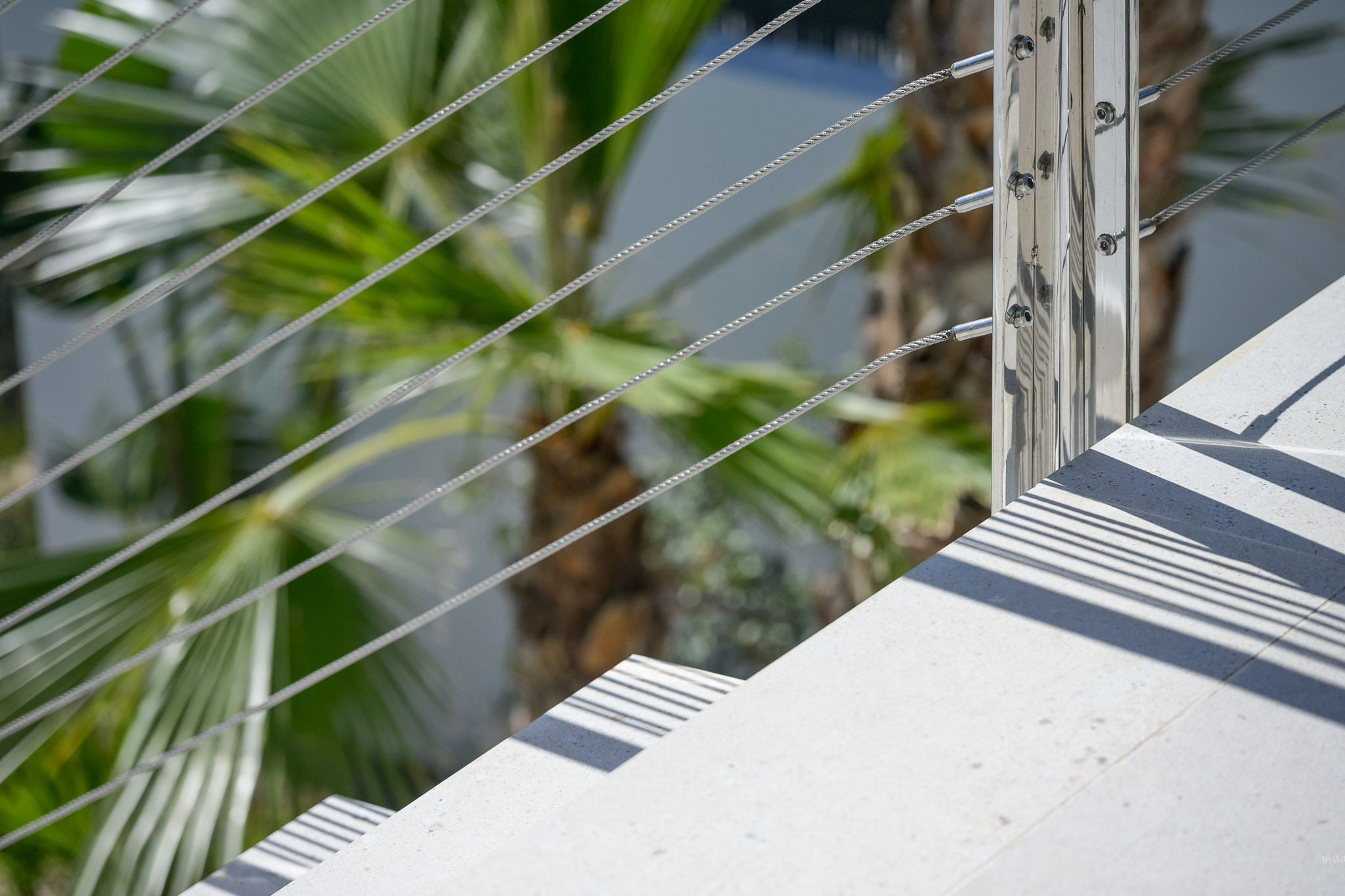 Detail van moderne buitentrap: lichtgekleurde treden, gepolijste metalen balustrade met staalkabels. Zonlicht werpt scherpe parallelle schaduwen, omringd door weelderig palmblad.