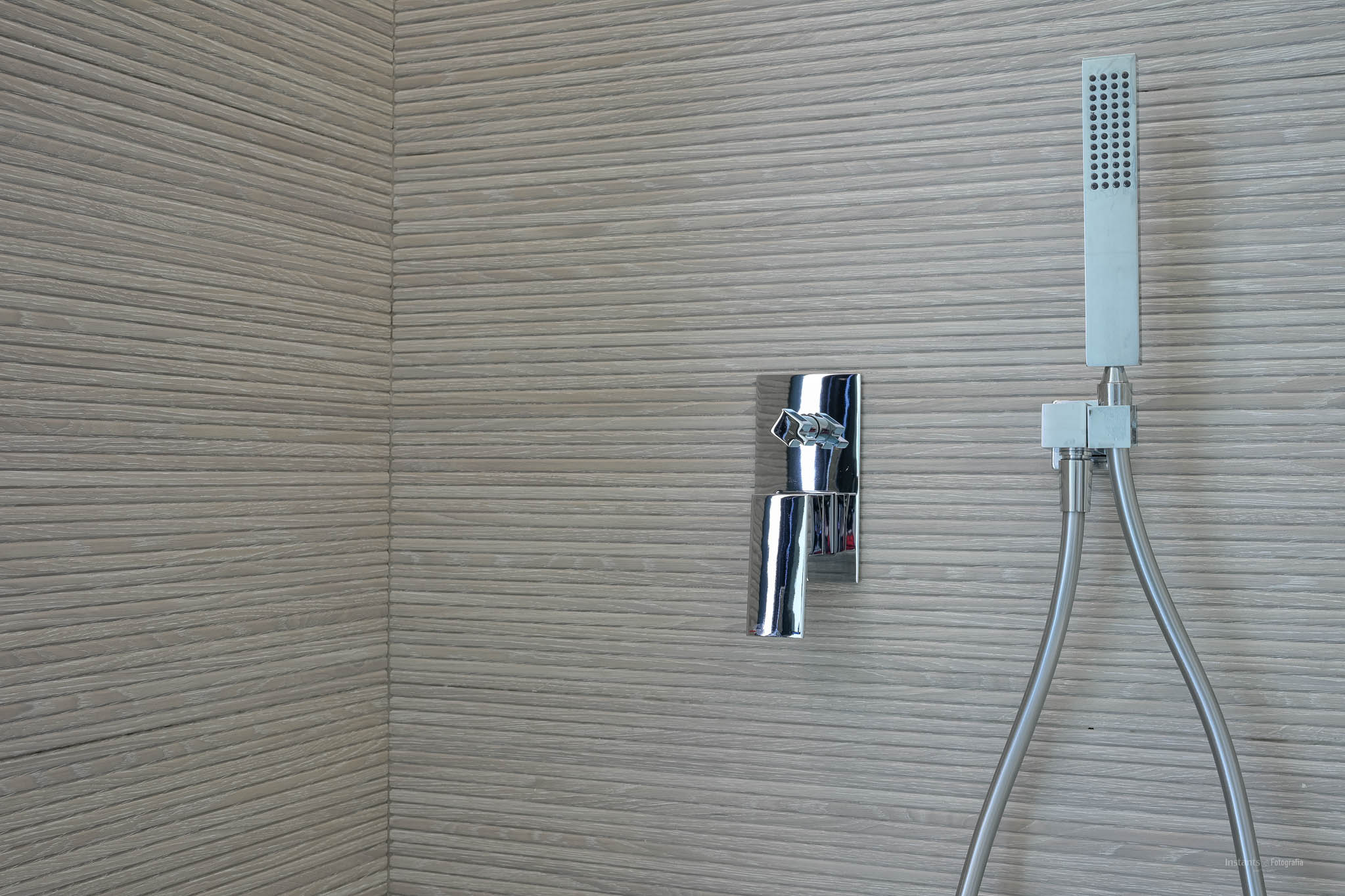 Douchehoek met chromen mengkraan en handdouche tegen een wand bekleed met horizontaal geribbelde textuurtegels.