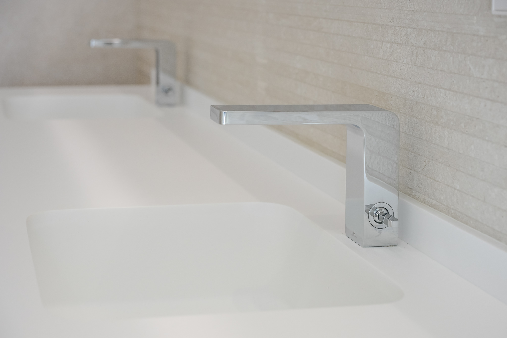 Dubbele wastafel met strakke chromen kranen en geïntegreerde witte wasbakken. De achterwand is bekleed met lichte, horizontaal gestructureerde tegels.
