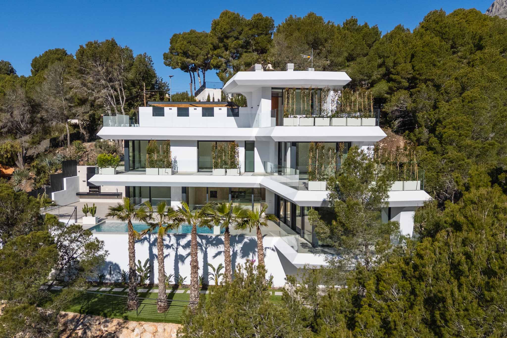 Hedendaagse villa met meerdere verdiepingen, witte gevel, ruime glazen terrassen, een zwembad met palmbomen en buitenkeuken, gelegen op een groene heuvel.