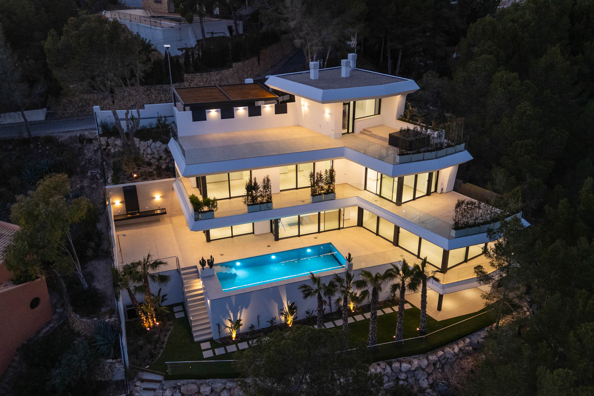 Luchtfoto van de moderne villa met meerdere verdiepingen in de schemering, met verlichte terrassen, het felblauwe zwembad en een buitenkeuken, elegant verlicht te midden van weelderig groen.