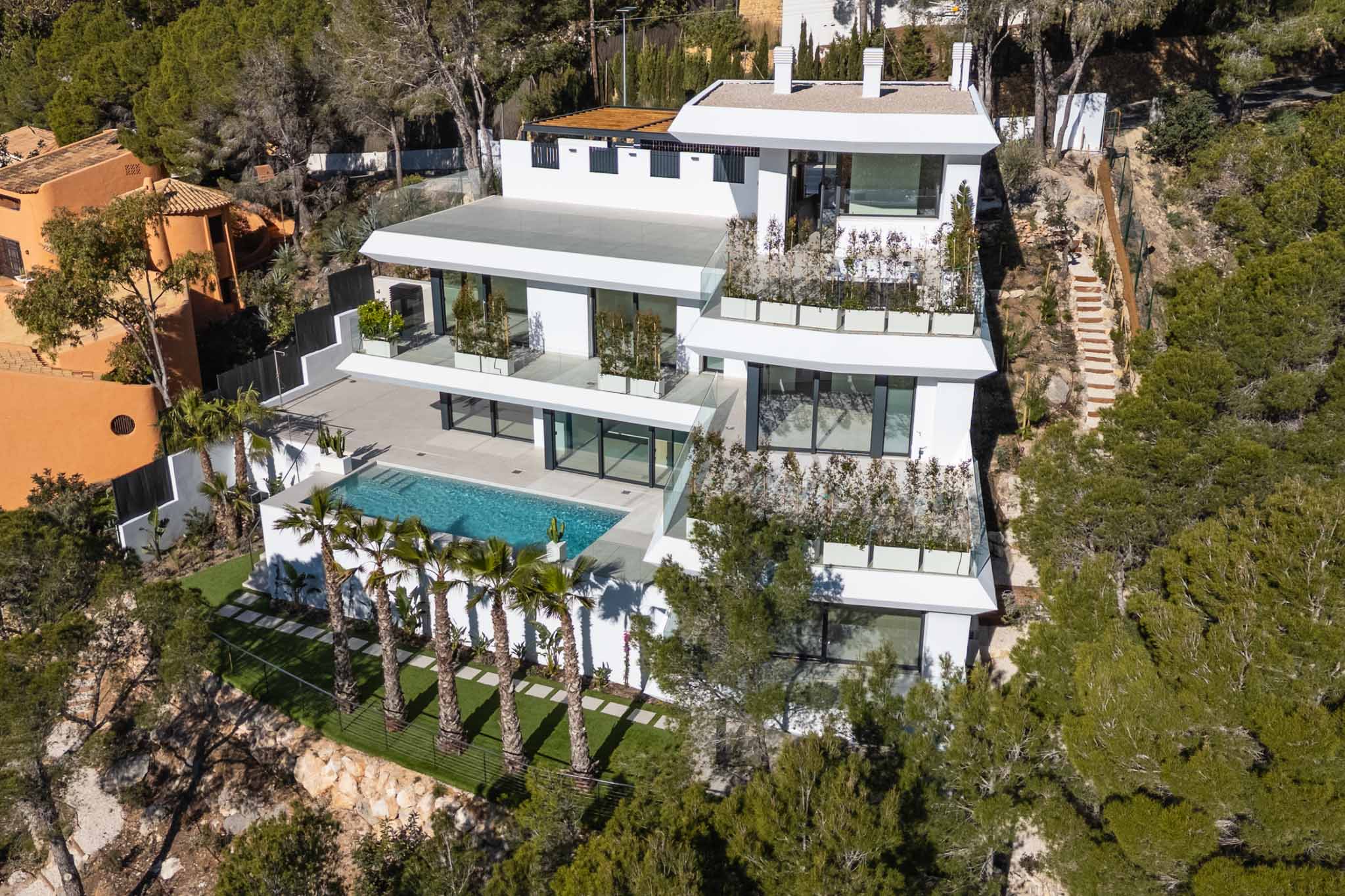 Luchtfoto van een moderne, meerlaagse witte villa, genesteld op een groene heuvel. Voorzien van een groot privézwembad, ruime terrassen met glazen balustrades en palmbomen, omgeven door mediterraan bos.