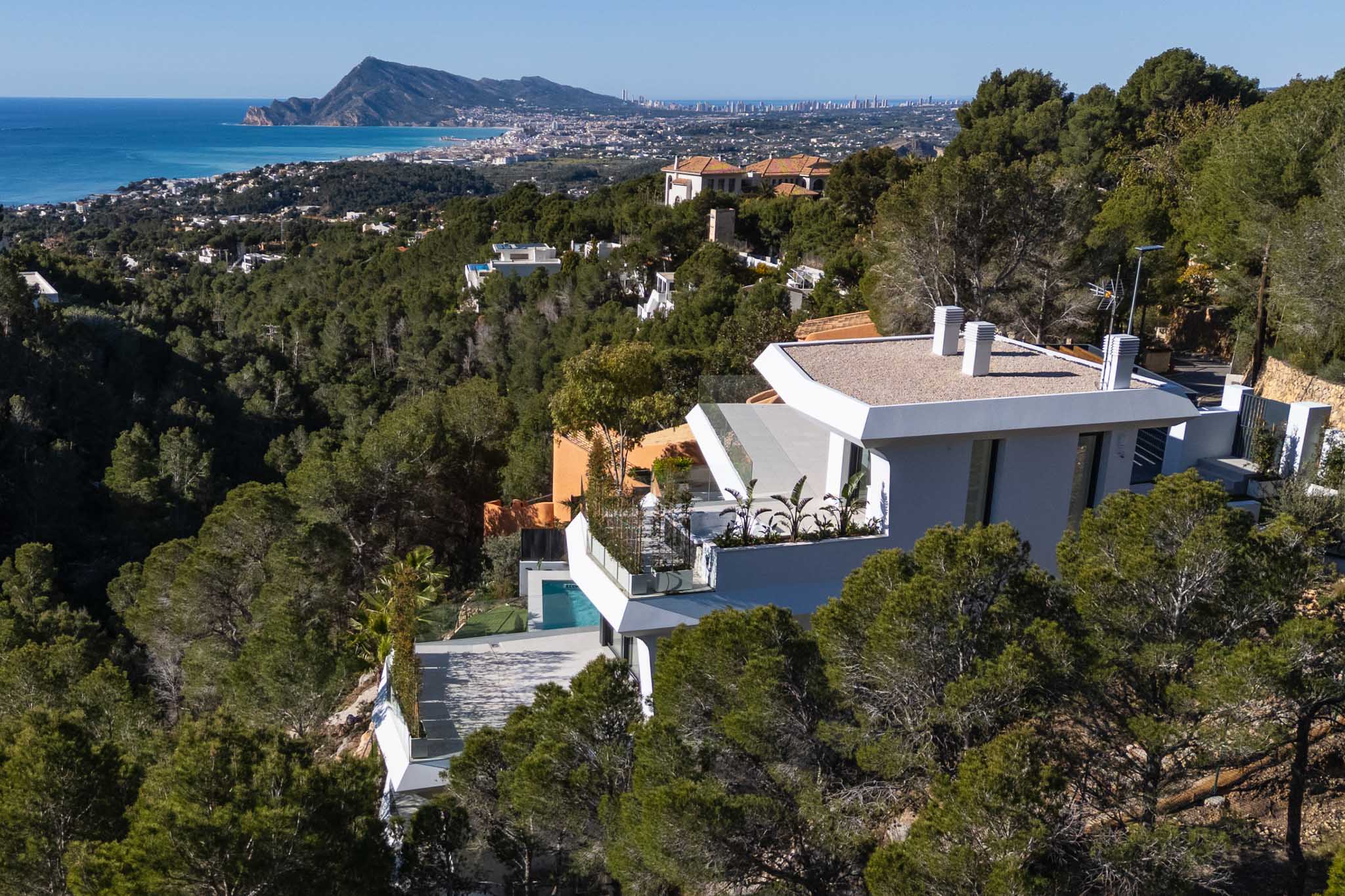 Luchtfoto van een moderne witte villa op een met pijnbomen begroeide heuvel, met panoramisch uitzicht op de Middellandse Zee en de kustlijn van Altea. Een privézwembad is zichtbaar op een lager terras.