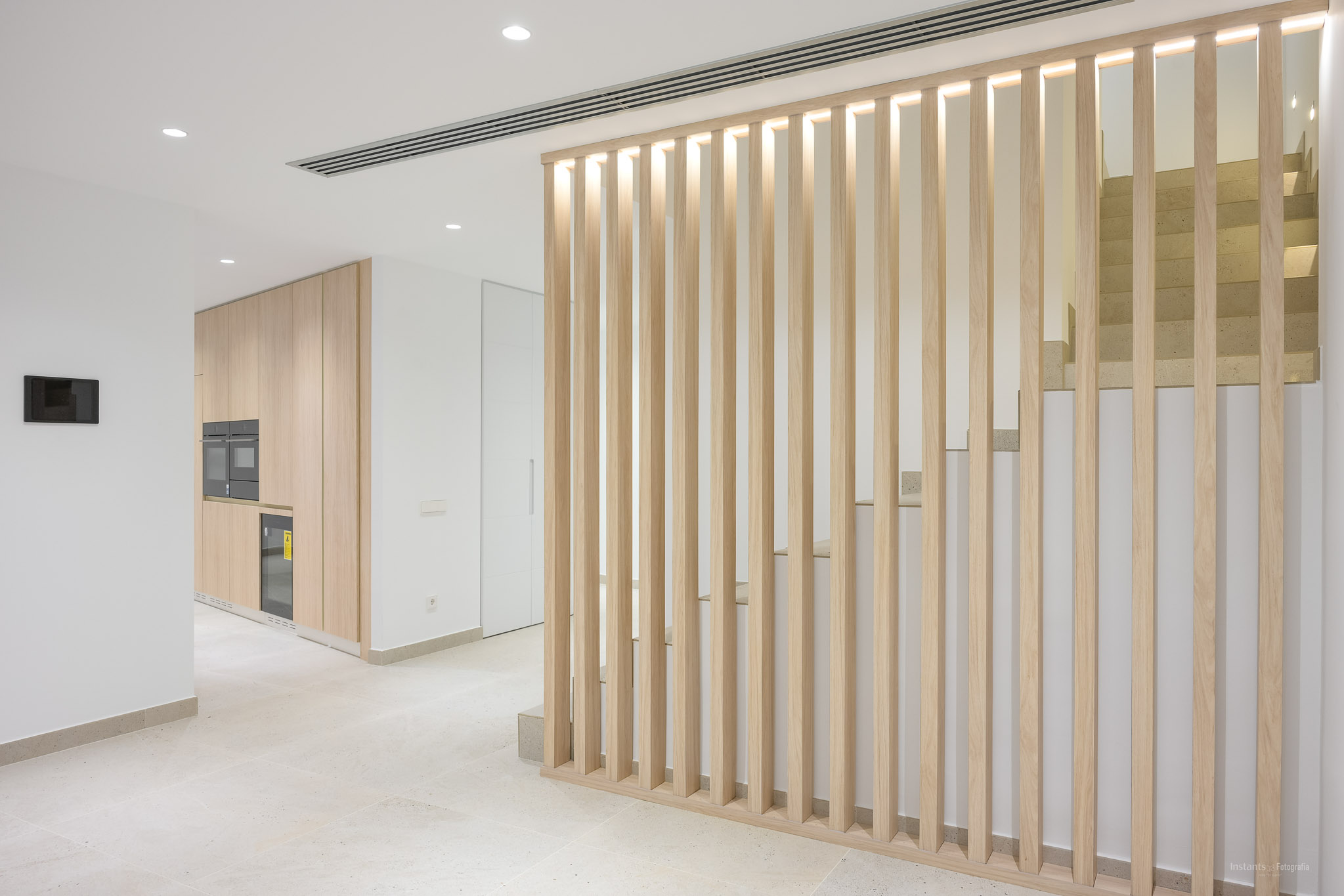 Modern interieur met een lichte houten lamellenwand en geïntegreerde verlichting, die een strakke keuken met inbouwapparatuur scheidt van een eigentijdse stenen trap.