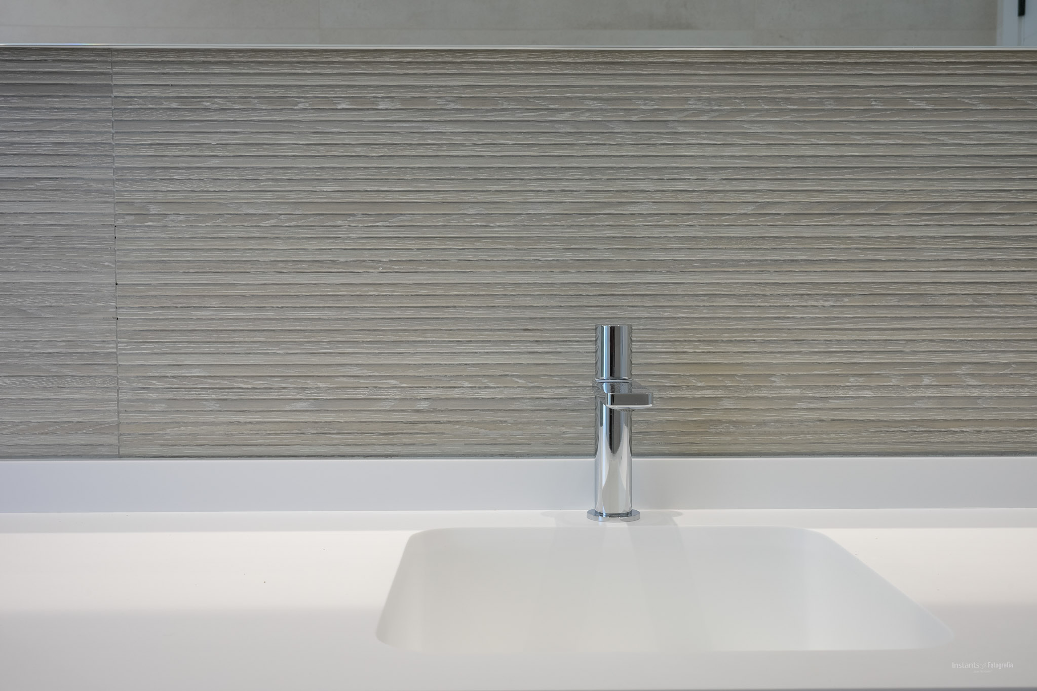 Modern wit geïntegreerd wastafelmeubel met strakke chromen kraan. De wand is bekleed met lichtgekleurde, horizontaal gestructureerde tegels, voor een minimalistische uitstraling.