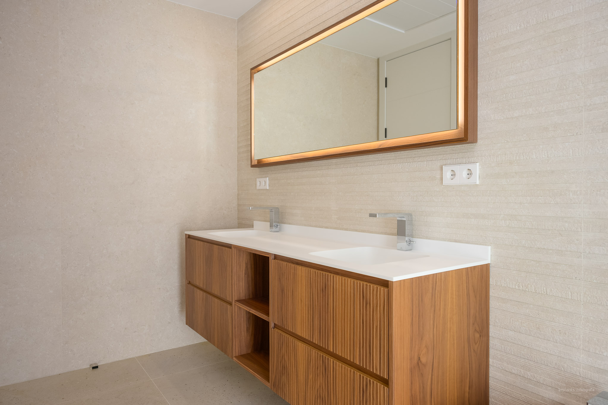 Modern zwevend houten badmeubel met dubbele wastafel en chromen kranen. Verlichte spiegel met houten lijst en getextureerde wandtegels.