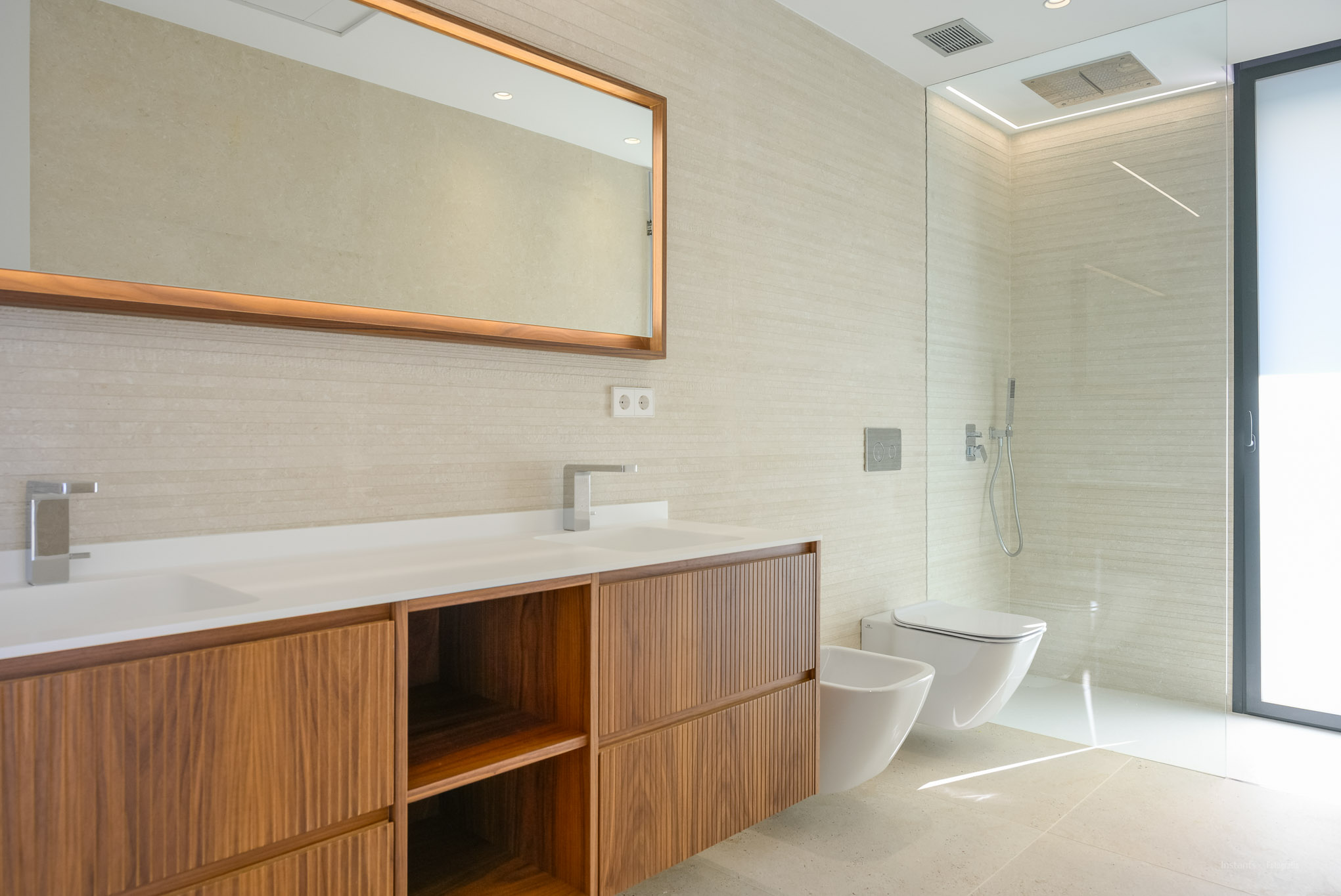 Moderne badkamer met dubbele wastafel, houten meubel en getextureerde wandtegels. Verlichte spiegel, inloopdouche, wandcloset en bidet zijn aanwezig.