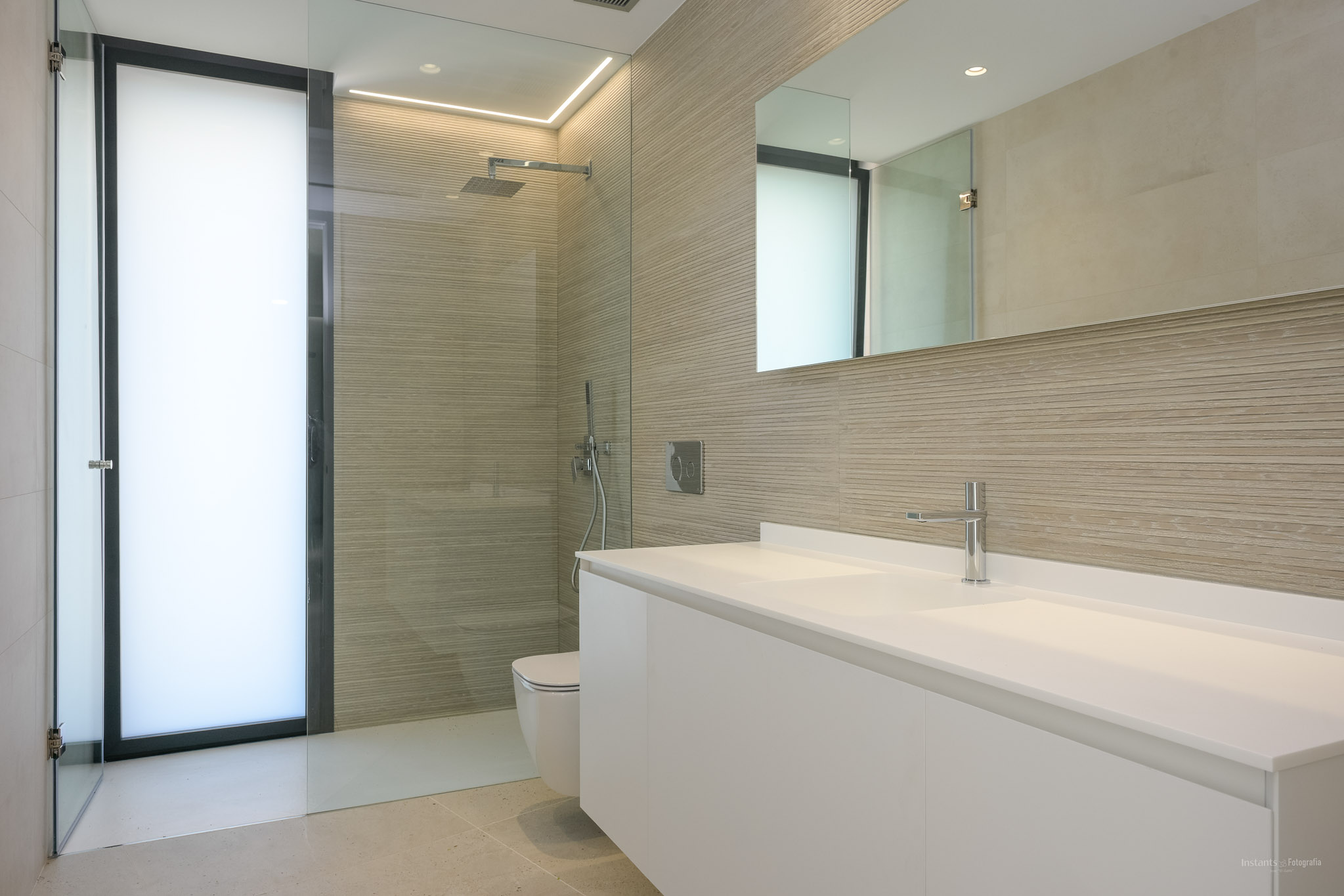 Moderne badkamer met dubbele zwevende witte wastafel, inloopdouche met getextureerde wandtegels en een matglazen deur, aangevuld met chromen kranen en geïntegreerde verlichting.