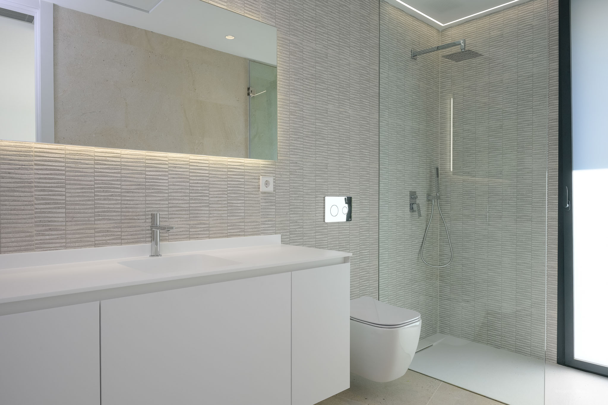 Moderne badkamer met wit wastafelmeubel, verlichte spiegel en getextureerde grijze wandtegels. Wandcloset en ruime inloopdouche met glazen scheidingswand.