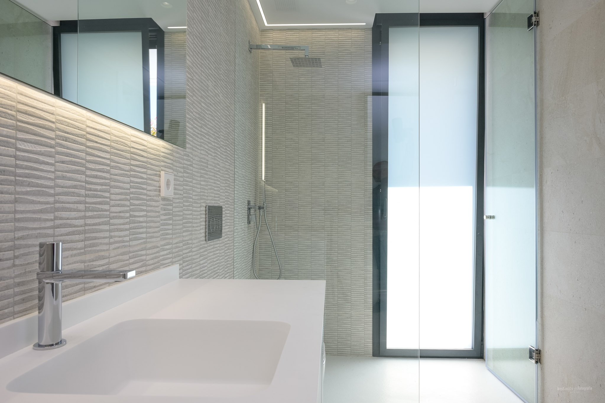 Moderne badkamer met witte wastafel, chromen kraan en getextureerde grijze wandtegels. Voorzien van een glazen douche met regendouchekop en LED-verlichte spiegel.
