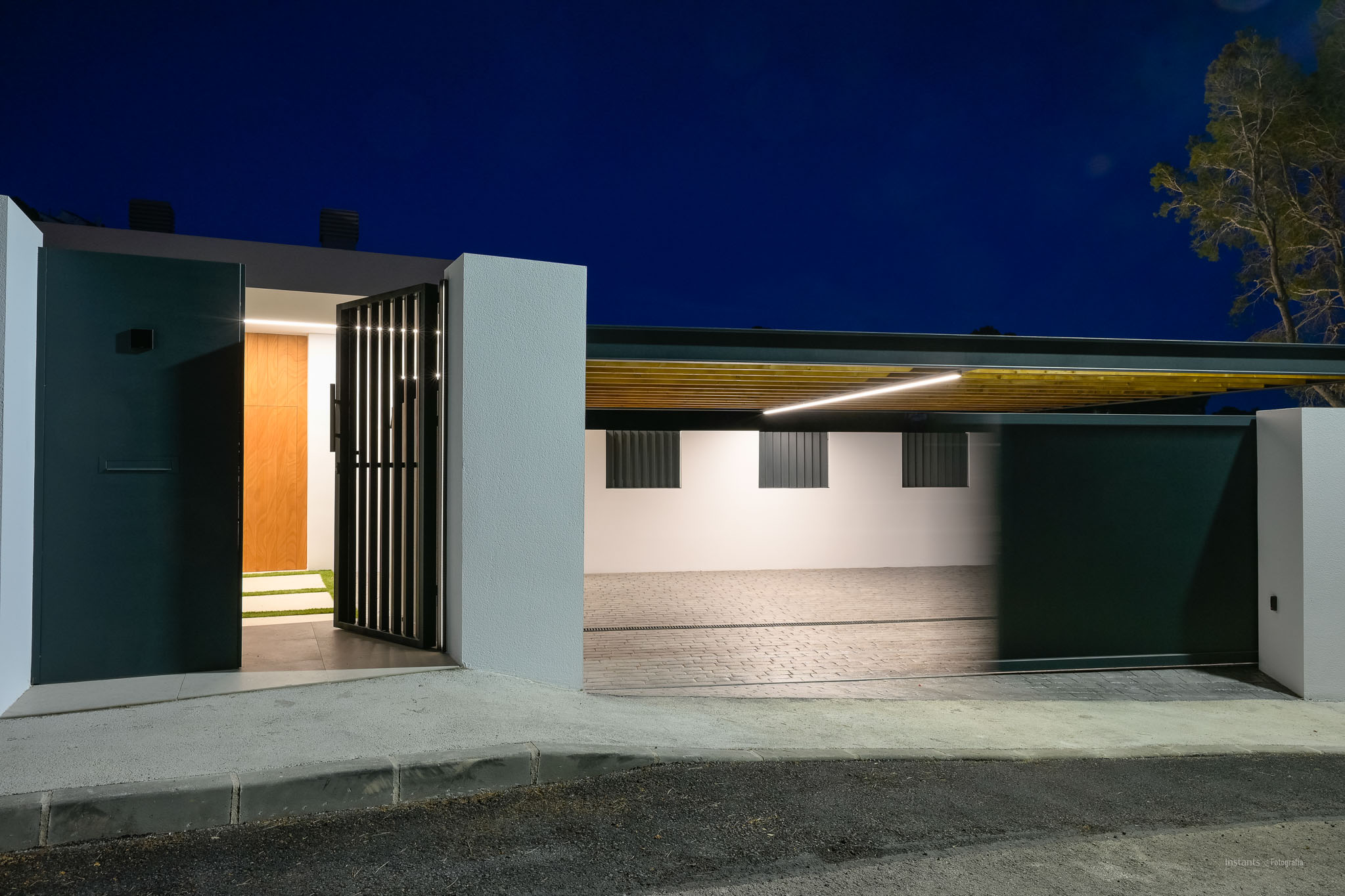 Moderne entree met donker voetgangershek en houten voordeur, naast een verlichte carport met lamellenplafond en lineaire LED-verlichting.