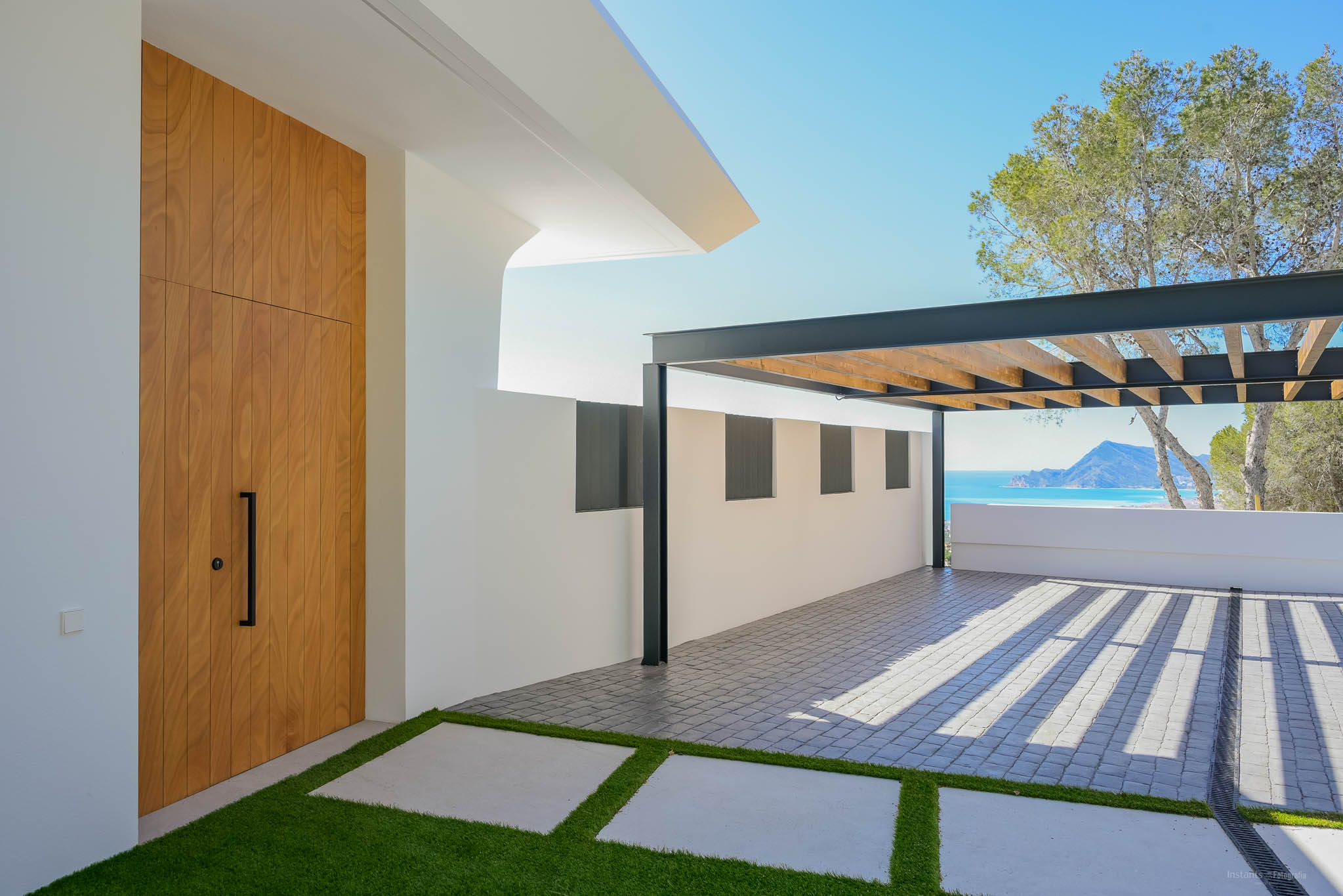Moderne entree met hoge houten deur, kunstgras pad en een bestrate parkeerplaats onder een metalen pergola. Vergezichten op de Middellandse Zee en bergen zijn zichtbaar.