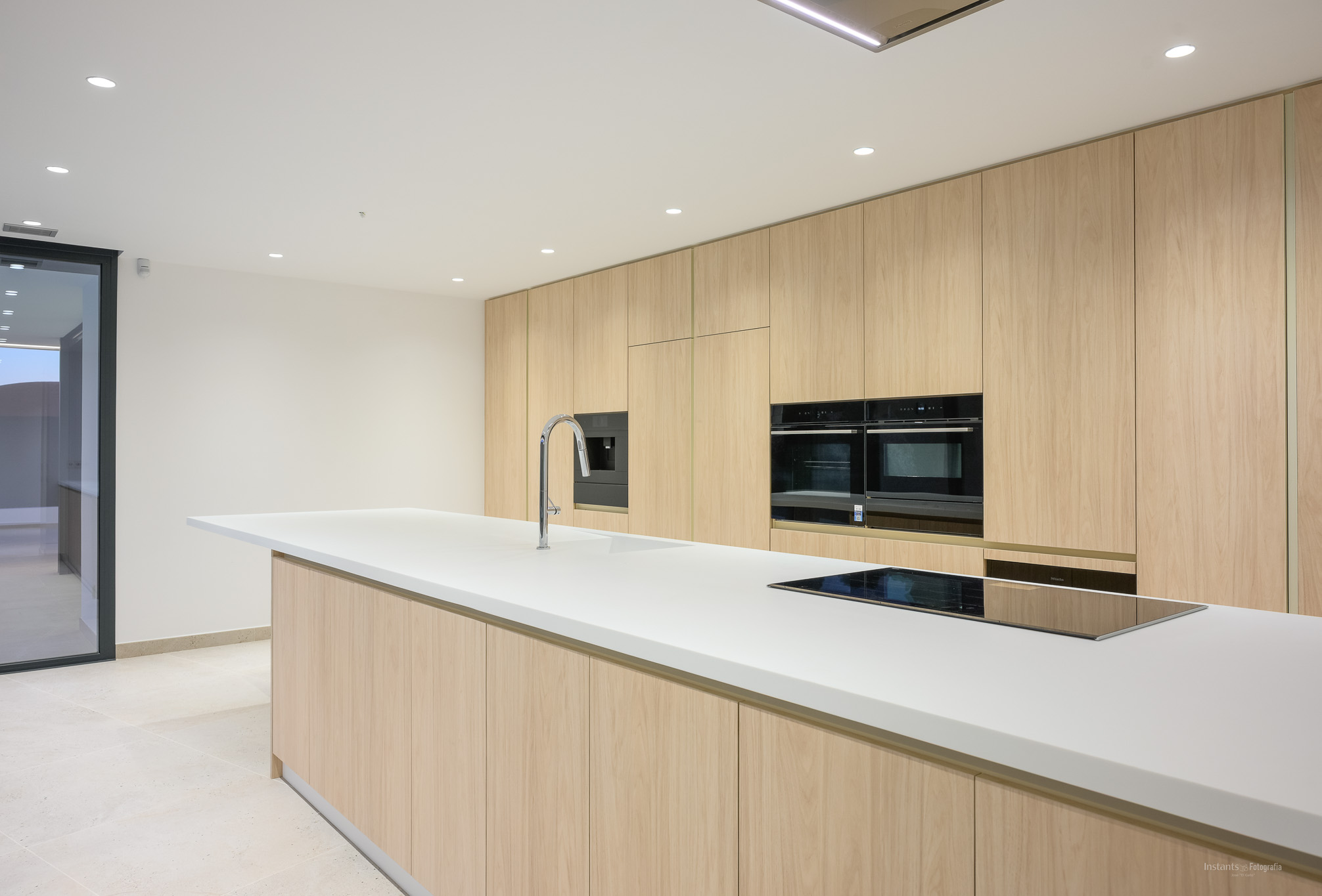 Moderne keuken met een groot wit eiland inclusief geïntegreerde spoelbak en inductiekookplaat, aangevuld met plafondhoge licht houten kasten en inbouwovens.