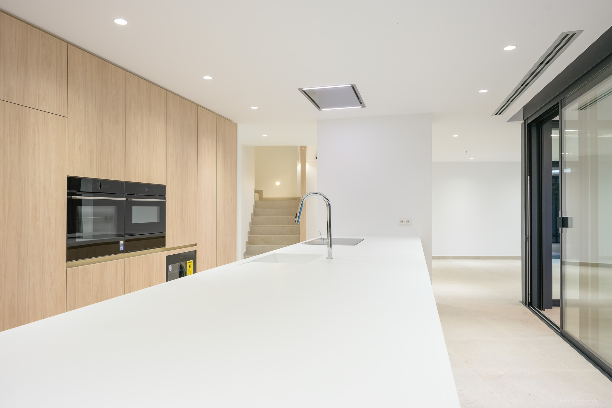 Moderne keuken met een lang wit eiland inclusief geïntegreerde spoelbak en chromen kraan, aangevuld met lichte houten kastenwand en ingebouwde dubbele ovens. Inbouwspots verlichten de eigentijdse ruimte.