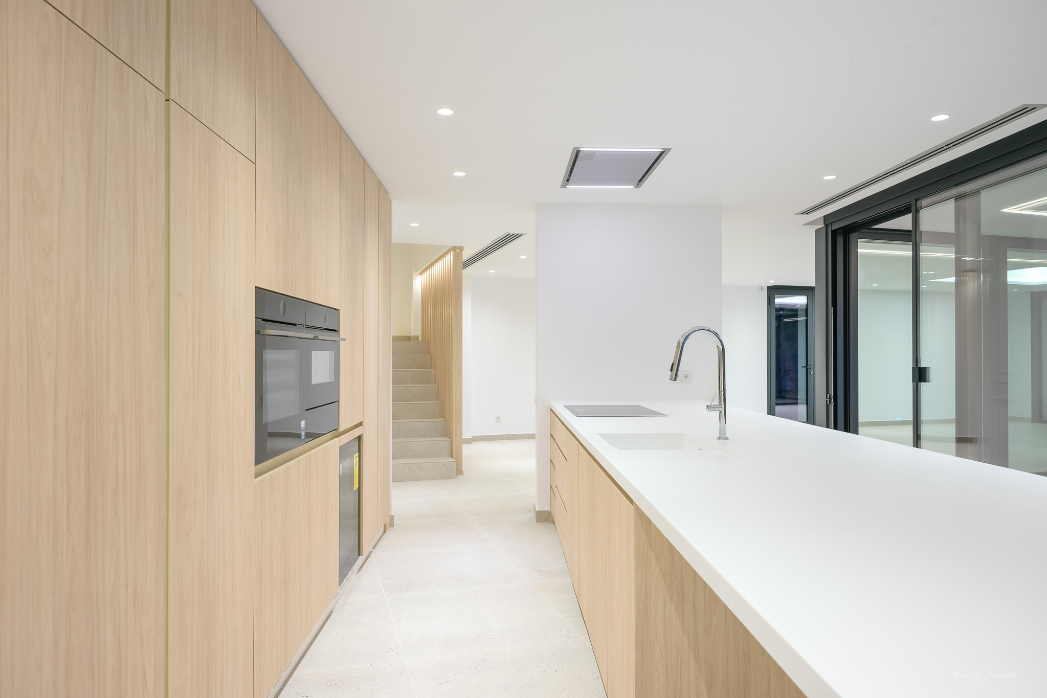 Moderne keuken met lichte houten kasten, wit werkblad en geïntegreerde apparatuur zoals een oven, inductiekookplaat en spoelbak met chromen kraan. Verbonden met trap en buitentoegang.