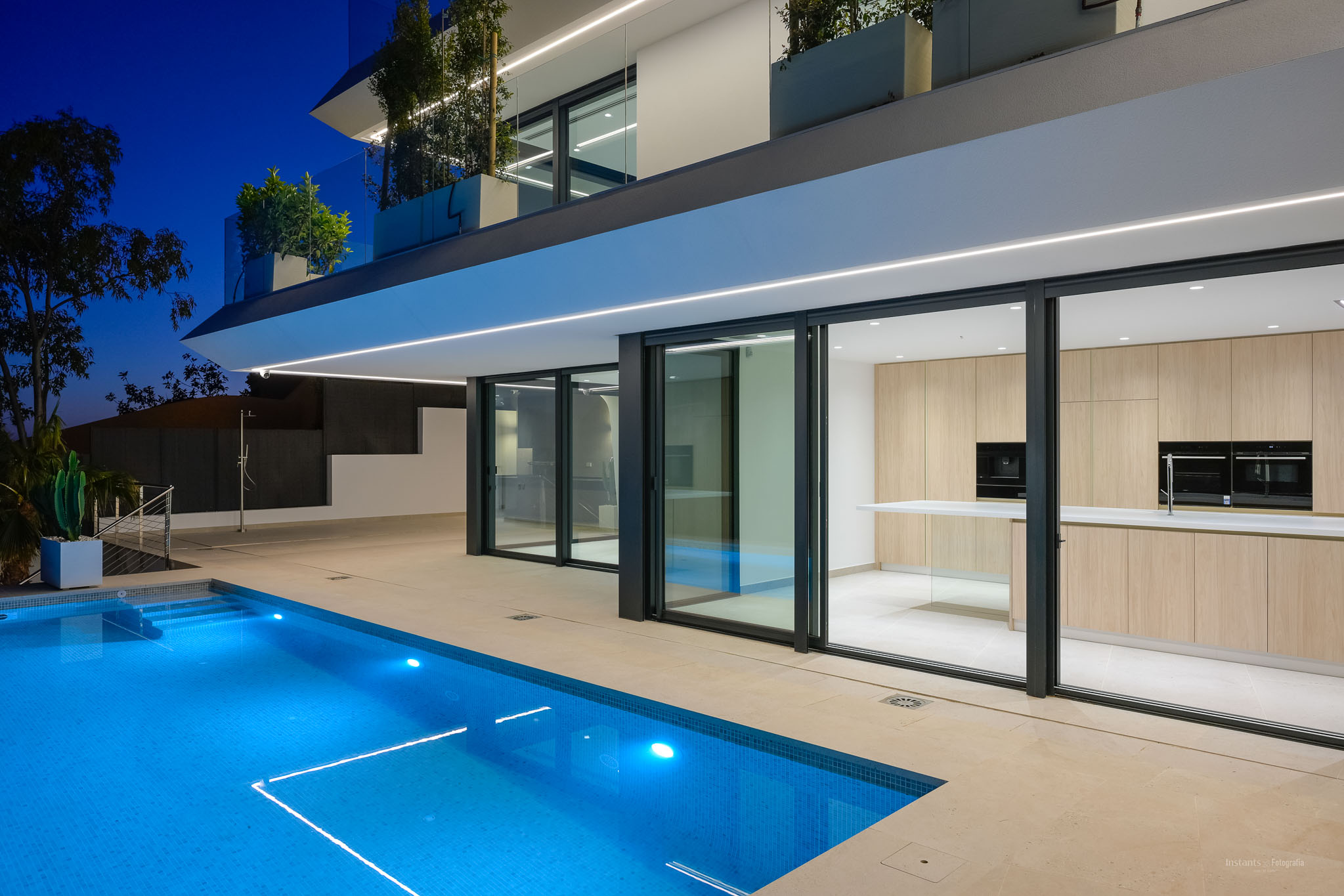 Nachtzicht op de moderne villa met verlicht blauw zwembad en strakke architectuur. Een lichte houten buitenkeuken is zichtbaar door de grote glazen schuifpuien.