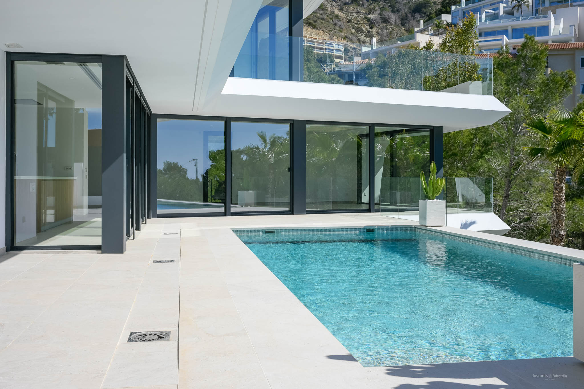 Privézwembad met blauwe mozaïektegels en lichtgekleurd stenen terras, geflankeerd door grote glazen schuifpuien naar het moderne interieur van de villa.