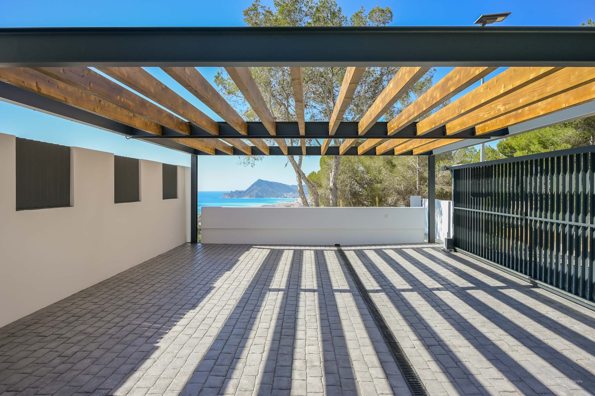 Ruime buitenruimte met pergola van houten balken en metalen frame op kasseien bestrating. Geniet van panoramisch zee- en bergzicht, met scherpe schaduwen van het natuurlijke licht.