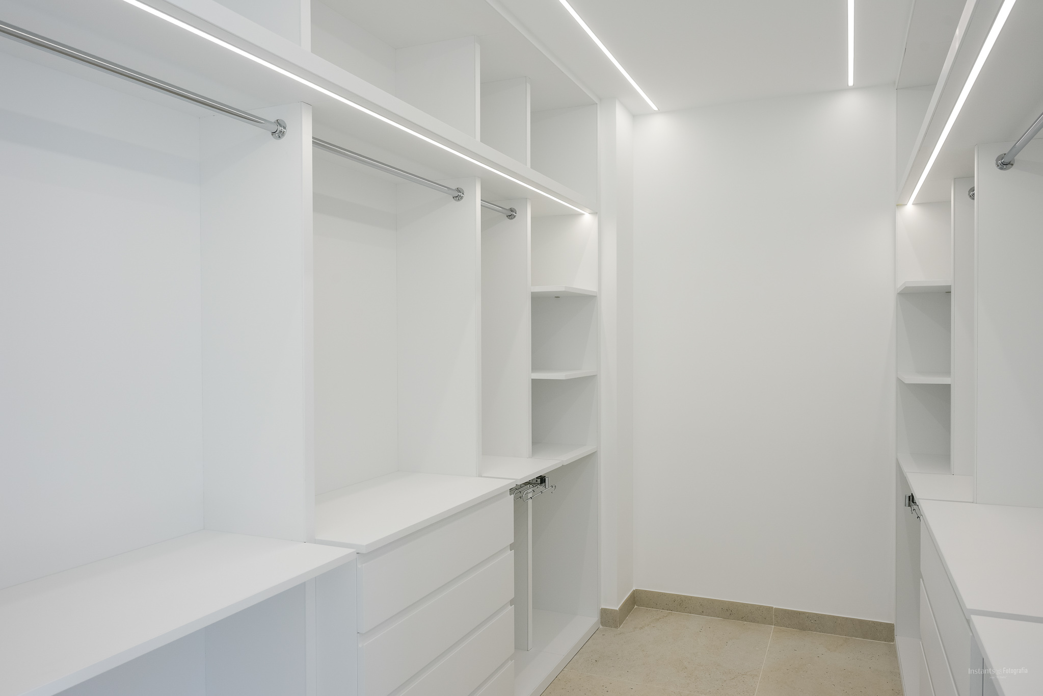 Ruime inloopkast met op maat gemaakte witte kasten, chromen kledingroedes en geïntegreerde LED-stripverlichting. Voorzien van diverse planken en lades, op een lichte tegelvloer.