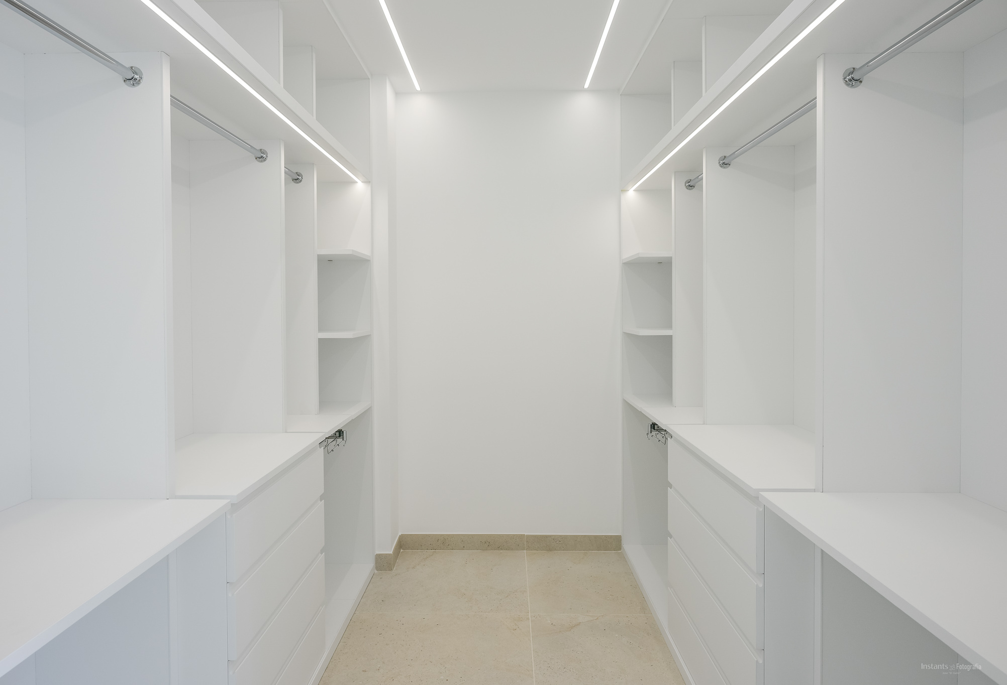 Ruime witte inloopkast met op maat gemaakte planken, laden en hangstangen, verlicht door geïntegreerde LED-strips en een lichte tegelvloer.