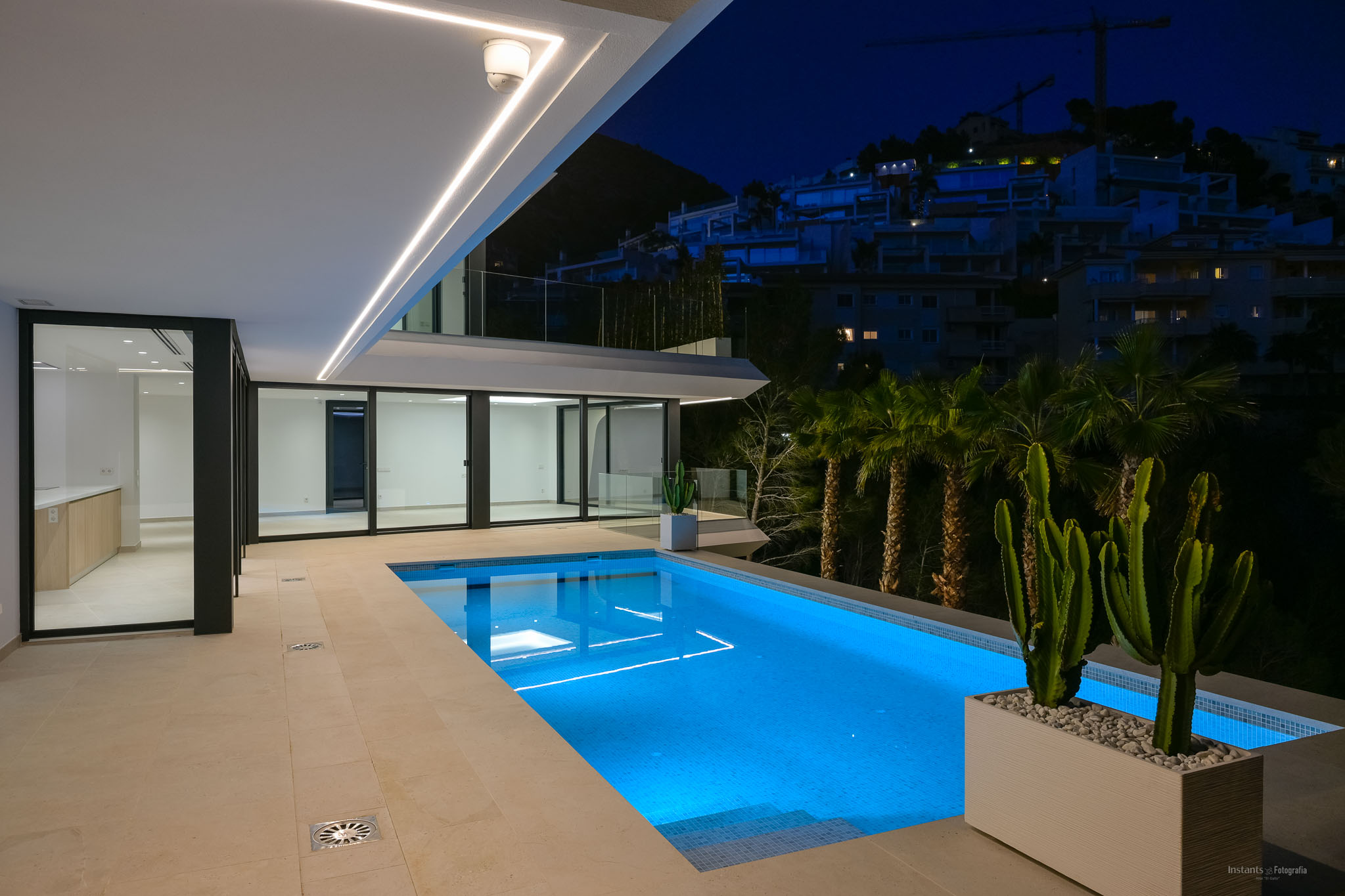 Verlicht zwembad 's nachts met levendig blauw onderwaterlicht en een ruim betegeld terras. Moderne lineaire plafondverlichting accentueert de architectuur, met uitzicht op een heuvel met verre gebouwen.