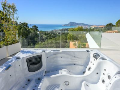 Buiten-jacuzzi op een betegeld terras met panoramisch uitzicht op de Middellandse Zee, de kustplaats en bergen. Glazen balustrade en groene beplanting creëren een open sfeer.