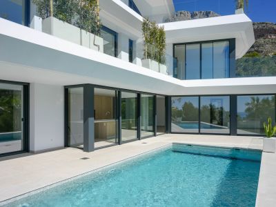 Buitenbad met lichte tegels en beige terras, geflankeerd door grote glazen puien die een buitenkeuken en het villa-interieur tonen, met uitzicht op de bergen.