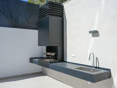 Buitenkeuken met geïntegreerde donkergrijze barbecue, een bijpassend aanrechtblad en een roestvrijstalen spoelbak. Een moderne chromen kraan completeert de lichte tegelwanden.