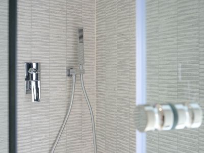 Detail van een moderne douche met grijze, gestructureerde rechthoekige tegels, een slanke chromen mengkraan en handdouche. Een glazen scherm is zichtbaar.