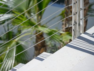 Detail van moderne buitentrap: lichtgekleurde treden, gepolijste metalen balustrade met staalkabels. Zonlicht werpt scherpe parallelle schaduwen, omringd door weelderig palmblad.