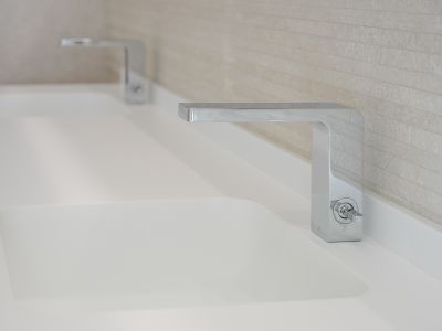 Dubbele wastafel met strakke chromen kranen en geïntegreerde witte wasbakken. De achterwand is bekleed met lichte, horizontaal gestructureerde tegels.