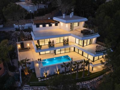 Luchtfoto van de moderne villa met meerdere verdiepingen in de schemering, met verlichte terrassen, het felblauwe zwembad en een buitenkeuken, elegant verlicht te midden van weelderig groen.