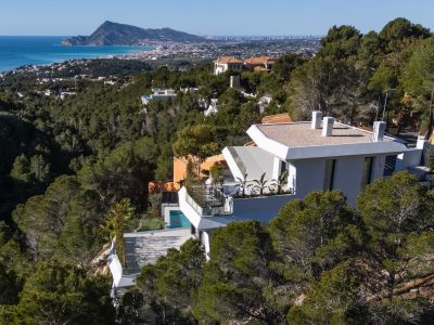 Luchtfoto van een moderne witte villa op een met pijnbomen begroeide heuvel, met panoramisch uitzicht op de Middellandse Zee en de kustlijn van Altea. Een privézwembad is zichtbaar op een lager terras.