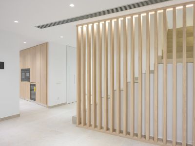 Modern interieur met een lichte houten lamellenwand en geïntegreerde verlichting, die een strakke keuken met inbouwapparatuur scheidt van een eigentijdse stenen trap.