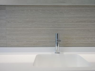 Modern wit geïntegreerd wastafelmeubel met strakke chromen kraan. De wand is bekleed met lichtgekleurde, horizontaal gestructureerde tegels, voor een minimalistische uitstraling.
