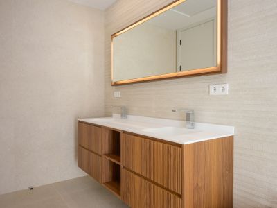 Modern zwevend houten badmeubel met dubbele wastafel en chromen kranen. Verlichte spiegel met houten lijst en getextureerde wandtegels.