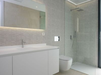 Moderne badkamer met wit wastafelmeubel, verlichte spiegel en getextureerde grijze wandtegels. Wandcloset en ruime inloopdouche met glazen scheidingswand.