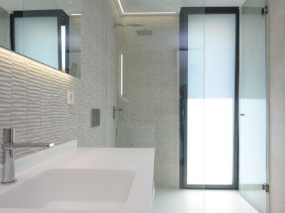Moderne badkamer met witte wastafel, chromen kraan en getextureerde grijze wandtegels. Voorzien van een glazen douche met regendouchekop en LED-verlichte spiegel.