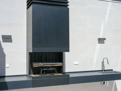 Moderne buitenkeuken met donkere geïntegreerde barbecue en strakke afzuigkap, bijpassend aanrecht met RVS spoelbak en chromen kraan, tegen een lichte tegelwand.