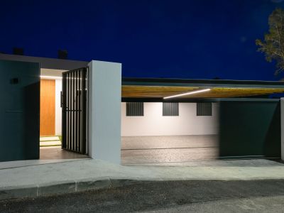 Moderne entree met donker voetgangershek en houten voordeur, naast een verlichte carport met lamellenplafond en lineaire LED-verlichting.