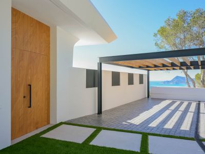 Moderne entree met hoge houten deur, kunstgras pad en een bestrate parkeerplaats onder een metalen pergola. Vergezichten op de Middellandse Zee en bergen zijn zichtbaar.