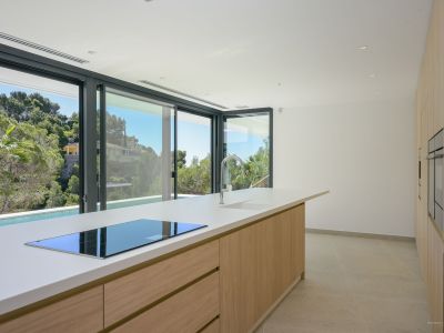 Moderne keuken met een groot eiland, wit aanrechtblad, geïntegreerde inductiekookplaat en chromen kraan. Schuifpuien bieden uitzicht op weelderige mediterrane begroeiing en een privézwembad.