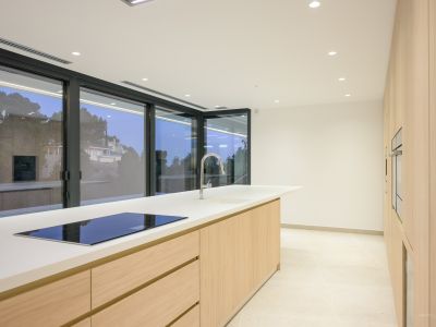 Moderne keuken met een groot kookeiland, wit werkblad en geïntegreerde inductiekookplaat. Lichte houten kasten en inbouwovens langs de wand, met grote schuifpuien.