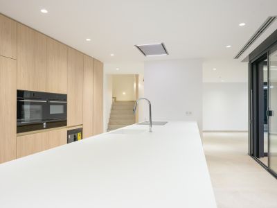 Moderne keuken met een lang wit eiland inclusief geïntegreerde spoelbak en chromen kraan, aangevuld met lichte houten kastenwand en ingebouwde dubbele ovens. Inbouwspots verlichten de eigentijdse ruimte.