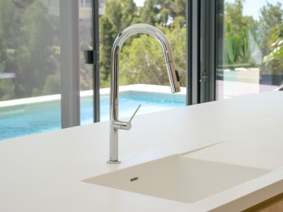 Moderne keuken met licht aanrechtblad en geïntegreerde spoelbak. Een chromen uittrekbare kraan kijkt uit op het zwembad en weelderig groen via grote glazen deuren.
