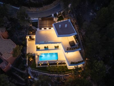 Nachtelijk luchtaanzicht van de moderne villa, met helder verlichte terrassen en het uitnodigende, blauwgloeiende zwembad. Meerdere buitenruimtes zijn zichtbaar, gelegen tegen de donkere, natuurlijke helling.