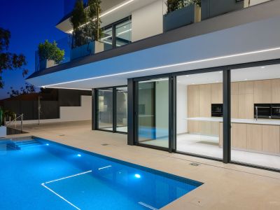 Nachtzicht op de moderne villa met verlicht blauw zwembad en strakke architectuur. Een lichte houten buitenkeuken is zichtbaar door de grote glazen schuifpuien.