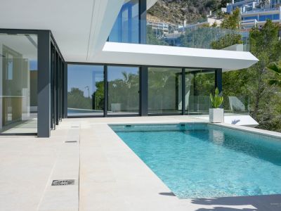 Privézwembad met blauwe mozaïektegels en lichtgekleurd stenen terras, geflankeerd door grote glazen schuifpuien naar het moderne interieur van de villa.