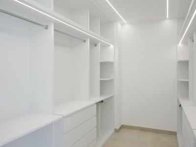 Ruime inloopkast met op maat gemaakte witte kasten, chromen kledingroedes en geïntegreerde LED-stripverlichting. Voorzien van diverse planken en lades, op een lichte tegelvloer.